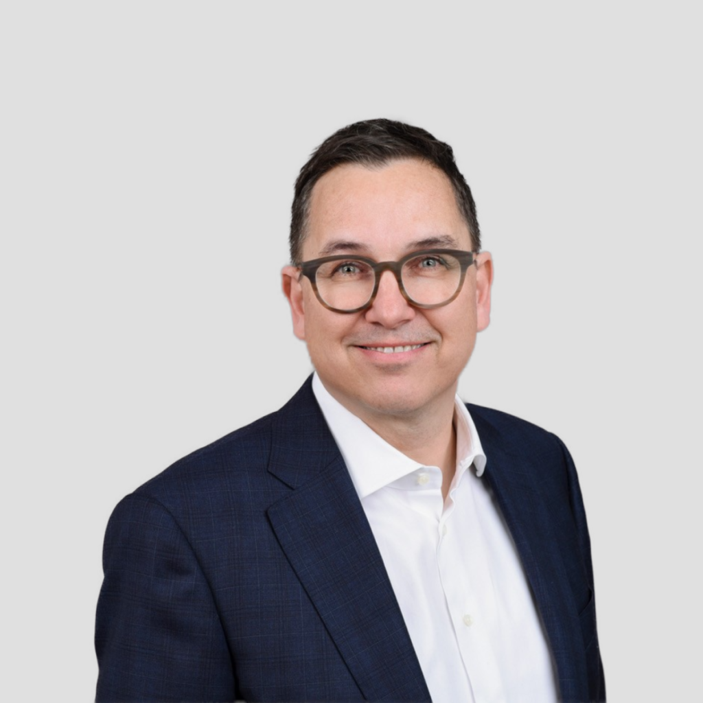 Professional headshot of a smiling man in a navy blazer and white shirt, wearing glasses, against a light gray background.